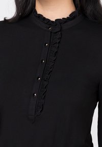 Haut noir avec un col volanté et une patte de boutonnage mi-longue avec quatre boutons métalliques. Tissu doux avec une texture lisse.