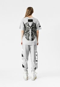 Camisa gris de gran tamaño con gráficos de alas negras en la parte posterior, combinada con pantalones jogger grises a juego que presentan diseños en blanco y negro, y botas blancas.