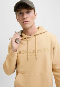 Sudadera beige con bolsillo canguro y cordones, con el texto en relieve "JACK&JONES PREMIUM", combinada con una gorra gris.
