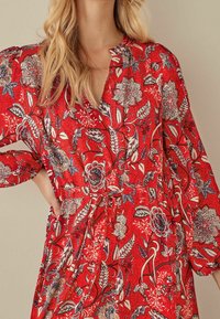 Robe portefeuille à fleurs rouges avec des manches longues, un décolleté en V et une taille à nouer. Le tissu présente une apparence douce et texturée avec des motifs complexes.