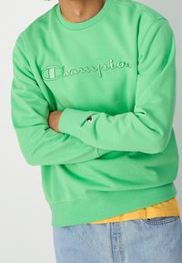 Person wearing a light green Champion sweatshirt with folded arms, layered over a mustard yellow shirt and light blue jeans.