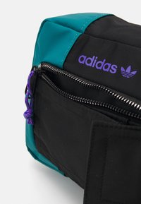 Svart och turkos Adidas-väska med lila logotyp, utrustad med en blixtlåsficka, texturerad tyg och en lila dragsko-accent.