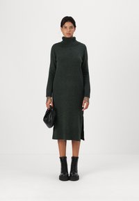 Abito lavorato a maglia di un verde scuro con colletto a imbuto, spacco laterale e polsini a costine; abbinato a stivali neri alla caviglia e una borsa nera.