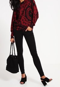 Donna che indossa una maglietta a maniche lunghe rossa e nera a fantasia, pantaloni skinny neri, sandali con tacco neri e tiene una borsa a tracolla nera.