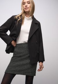 Musta, villasekoitusmateriaalia oleva takki, jossa on kärkipäälaki, nappikiinnitys ja valkoisen poolopaidan päällä. Malli yhdistää sen teksturoidun harmaan minihameen kanssa.