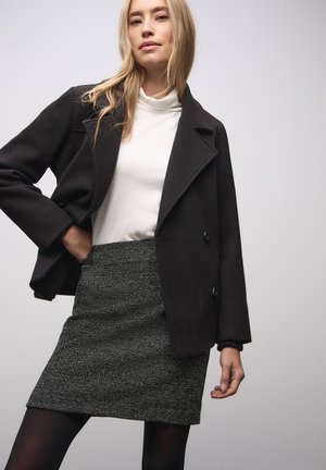 Schwarze Jacke aus Wollmischung mit spitzen Reversknöpfen und Knopfverschluss, getragen über einem weißen Rollkragenpullover. Das Modell kombiniert sie mit einem strukturierten grauen Minirock.