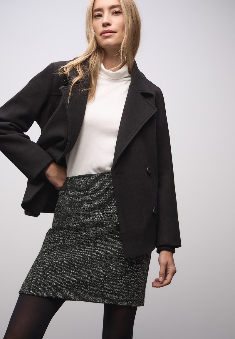 Musta, villasekoitusmateriaalia oleva takki, jossa on kärkipäälaki, nappikiinnitys ja valkoisen poolopaidan päällä. Malli yhdistää sen teksturoidun harmaan minihameen kanssa.