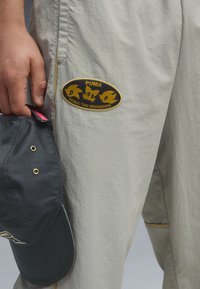 Beige bukser lavet af letvægtsstof med et sort og gult Sonic the Hedgehog-patch. En sort kasket med guld accenter ligger i nærheden.