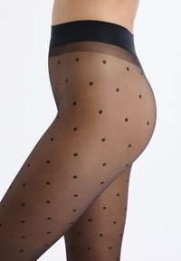 Primo piano di una persona che indossa collant neri trasparenti con piccoli pois neri e una fascia in vita nera solida su sfondo bianco.