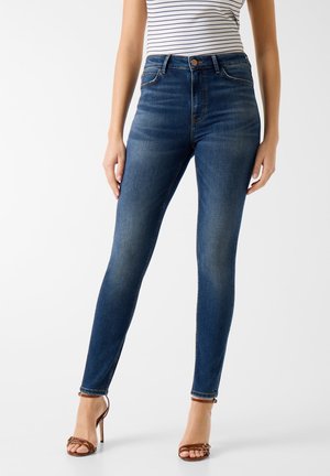 Kobieta w ciemnoniebieskich, obcisłych dżinsach z wysokim stanem, w parze z paskowanym, bezrękawnym topem i brązowymi sandałami na wysokim obcasie.