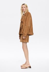 Tan nauwsluitende oversized jas en bijpassende shorts, gemaakt van een zacht materiaal. Tan sandalen hebben een decoratieve accent en een laag blokhak.