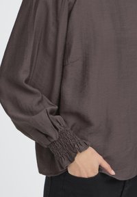 Brun langærmet bluse med en tekstureret finish, der har samlede manchetter og en afslappet pasform. Viser en sidelomme og et glat stoffald.