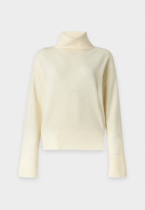 Femme debout portant un pull à col roulé crème, un pantalon noir raccourci et des chaussures plates noires sur un fond blanc uni.