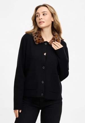 Frau trägt eine schwarze, mit Knöpfen versehene Strickjacke mit braunem Leopardenmuster aus Kunstfell als Kragen und schaut seitlich vor schlichtem Hintergrund.