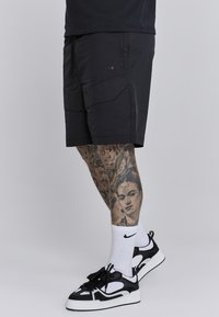 Mustat urheilushortsit, joissa on vetoketjulliset taskut ja kulmikkaat paneelit. Mallilla on päällä valkoiset Nike-sukat ja musta-valkoiset lenkkarit.