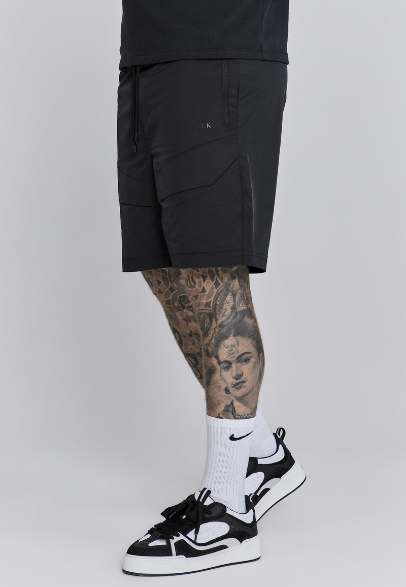 Mustat urheilushortsit, joissa on vetoketjulliset taskut ja kulmikkaat paneelit. Mallilla on päällä valkoiset Nike-sukat ja musta-valkoiset lenkkarit.