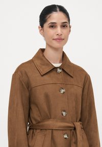 Braune Wildlederjacke mit Kragen, vier Knöpfen, tailliertem Gürtel und zwei seitlichen Taschen, mit einer glatten Textur.
