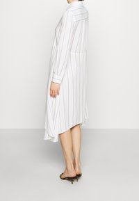 Abito camicia bianco con righe verticali nere, maniche lunghe e orlo curvo. Realizzato in tessuto leggero con una vestibilità comoda.