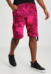 Rosa shorts med tropiskt mönster och svarta detaljer, med justerbara dragsnören, tillverkade av lättviktsmaterial, kombinerade med svarta slip-on skor.