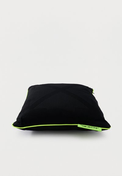 Coussin carré noir avec passepoil vert néon et une petite étiquette verte néon portant l'inscription "OBJECTS" sur un fond blanc uni.