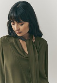 Olive green blouse with a soft, draped neckline and a tied detail at the collar. Smooth texture, long sleeves, and a relaxed fit.