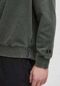 Grauer Sweatshirt aus strukturiertem Stoff. Auffällige Merkmale sind der gerippte Saum, gestickte Akzente und "ARC" auf dem Ärmel gestickt.