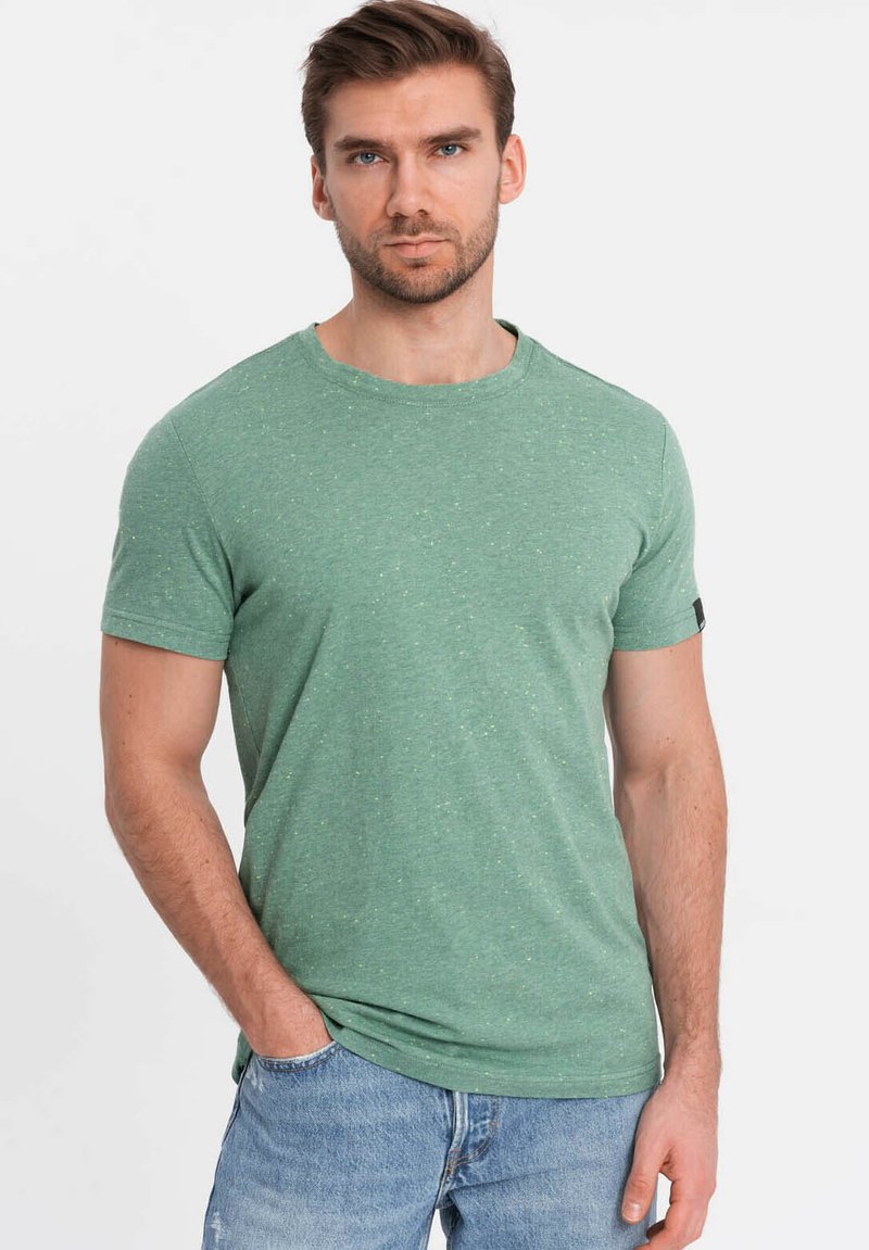 Camiseta verde de manga corta hecha de una tela suave con un sutil patrón moteado. Escote redondo con una etiqueta negra en la manga izquierda.