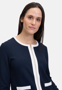 Veste navy avec des accents blancs, fermeture éclair et tissu texturé. Comprend deux poches avant et un col rond.