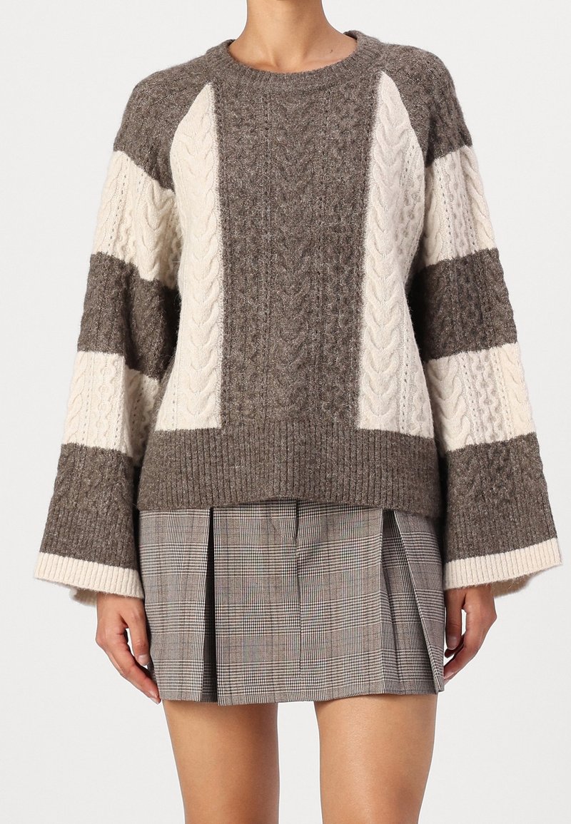 Femme portant un pull torsadé marron et crème avec des manches larges et une mini-jupe plissée à carreaux marron.