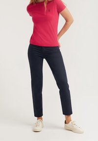 Žena v jasně růžovém tričku s krátkým rukávem, tmavě modrých úzkých kalhotách a bílých teniskách, stojí s jednou rukou na boku.