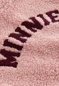 Strukturiert pinker Stoff mit einer plüschigen, flauschigen Oberfläche und maroonfarbenem Textdetails; zeichnet sich durch ein auffälliges, kontrastierendes Schriftbild aus.