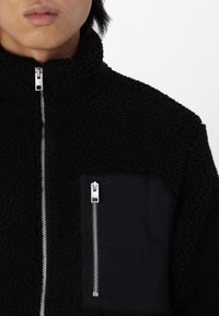 Schwarze Fleece-Jacke mit strukturiertem Oberflächenmaterial, versehen mit einem Reißverschluss und einer kontrastierenden schwarzen Stofftasche mit Reißverschlussdetail.
