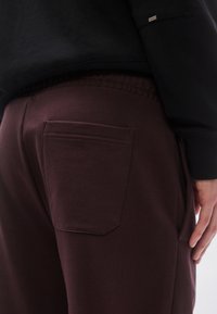 Pantaloni de trening burgund cu o textură fină, prevăzuți cu un buzunar pe spate și talie elastică. Asortați cu un hanorac negru, parțial vizibil.