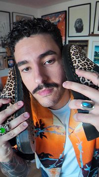 Asmenybės laikomos leopardinės rašto bateliai su juodais guminiais padais ir tekstūruotomis šoninėmis. Fone matoma spalvingų raštų marškiniai.