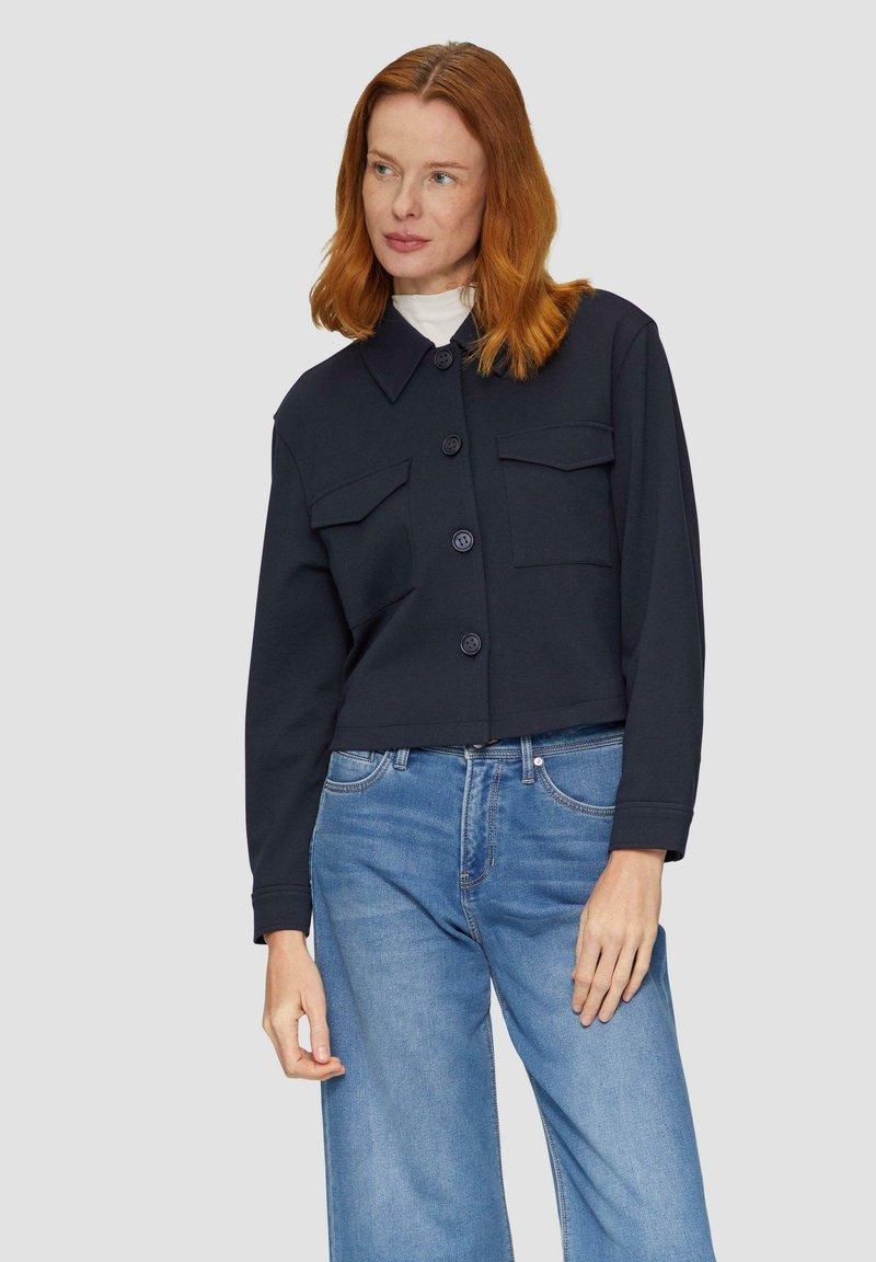 Donkerblauwe cropped jas met een kraag, sluiting met knopen aan de voorkant en twee borstzakken; gedragen boven een lichte blauwe wijde jeans.