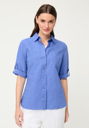 Woman wearing a light blue button-up shirt with rolled sleeves and white pants, standing against a plain light background.