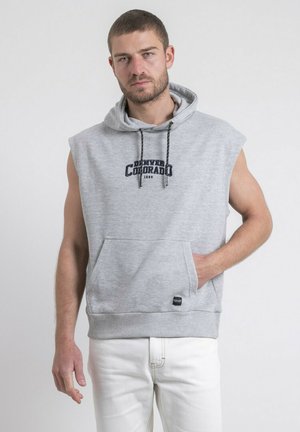 Homme portant un sweat à capuche gris sans manches avec le texte « Denver Colorado » et un pantalon blanc, debout avec une main dans la poche du sweat.