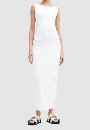 Robe maxi blanche sans manches avec une coupe décontractée, présentant un décolleté arrondi et un détail froncé sur le côté, associée à des sandales plates avec de grandes boucles.