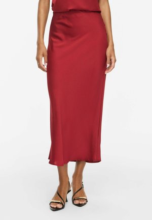 Mannequin portant une jupe midi rouge avec un haut assorti et des sandales noires à brides, sur un fond blanc uni.