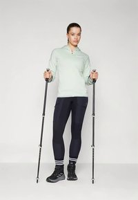 Νεαρή γυναίκα στέκεται κρατώντας δύο μαύρες ραβδώσεις πεζοπορίας, φορώντας ανοιχτό πράσινη φούτερ με φερμουάρ, μαύρα κολάν, ριγέ κάλτσες και μαύρα παπούτσια πεζοπορίας.