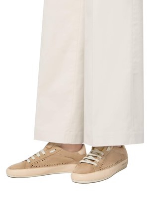 Sneakers in suede beige con design perforato, suola piatta in gomma crema e lacci bianchi a contrasto, abbinati a pantaloni larghi bianchi.