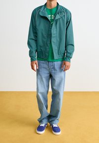 Green nylon jacket with snap buttons layered over a green t-shirt, paired with light blue jeans and blue suede shoes on a yellow floor.