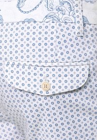Tissu blanc avec des motifs floraux et géométriques bleus. Possède une poche à rabat boutonnée avec un bouton crème. Matériau texturé et léger.