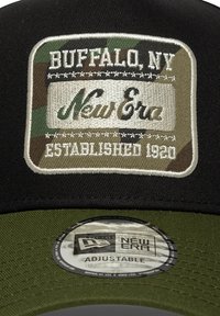 Čierna nastaviteľná čiapka s maskáčovým nášivom, na ktorom je vyšité "Buffalo, NY" a "New Era" v bielej farbe, a okrúhla nálepka New Era pod tým.