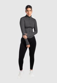 Tæt siddende mørkegrå zip-up top med tommelhuller, parret med sorte leggings. Model iført hvide sneakers, stående mod en lys grå baggrund.