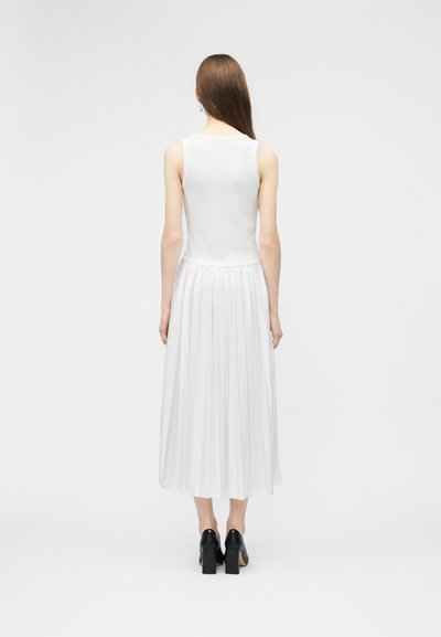 Femme aux longs cheveux bruns portant un haut sans manches blanc et une jupe midi plissée blanche, debout, de dos, sur un fond uni.