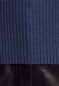 Pletený svetr v tmavě modré barvě s vertikální žebrovanou strukturou, doplněný hladkým černým koženým spodním lemem. Bez ozdob nebo vzorů.