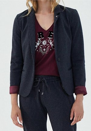 Nainen, jolla on tummansiniraidallinen bleiseri ja housut sekä viininpunainen kuvioitu T-paita, jossa on luurankoja ja taivaankappaleiden kuvioita.