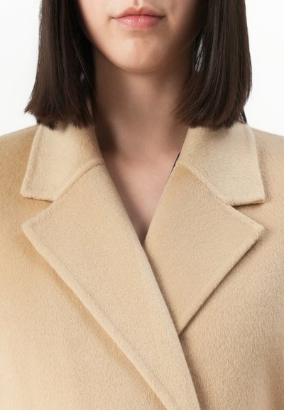 Nära bild av en kvinna som bär en beige ullrock med breda slag och rakt, mörkbrunt hår som når precis ovanför axlarna.