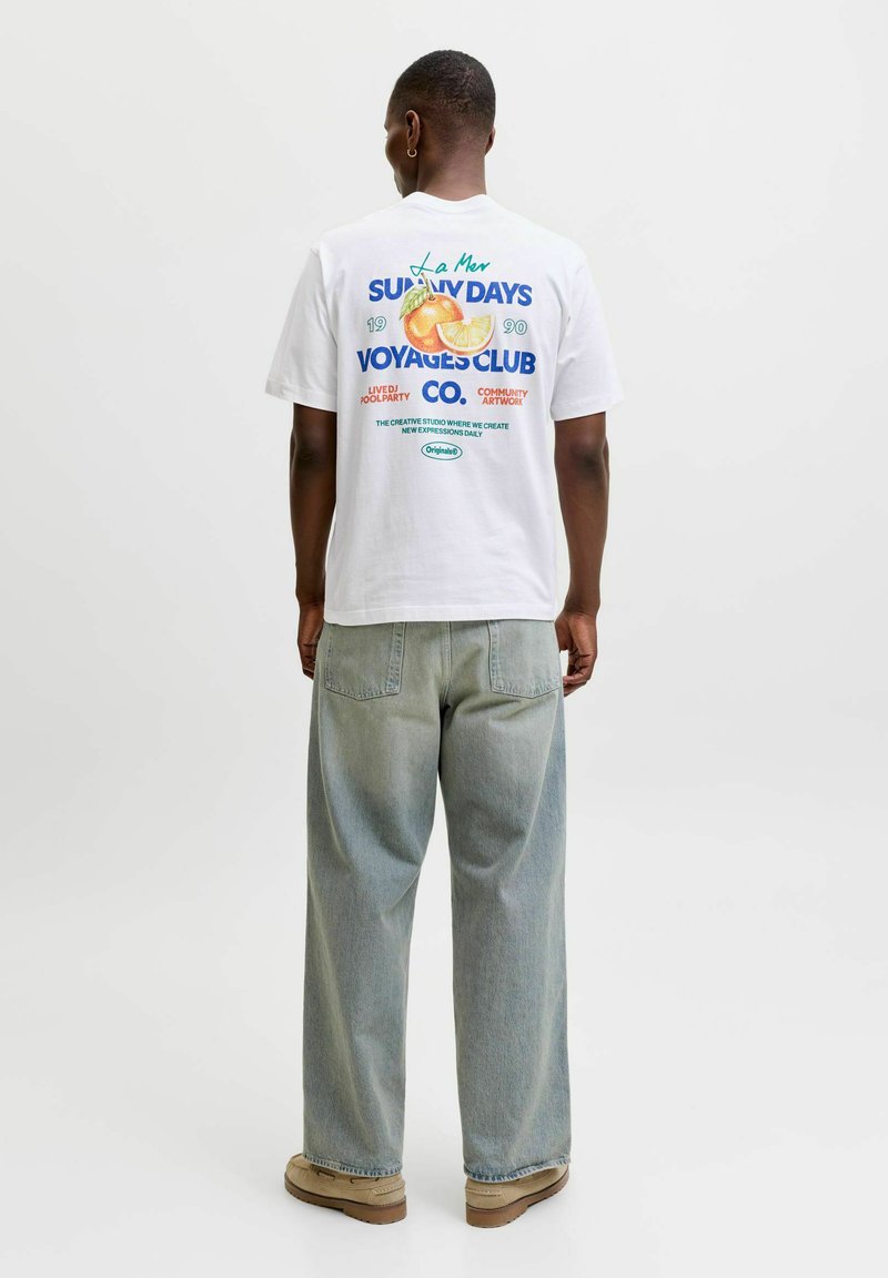 Άνδρας που φοράει λευκό t-shirt με πορτοκαλί σχέδιο και κείμενο, φαρδιά ανοιχτό μπλε τζιν, μπεζ παπούτσια, στέκεται με την πλάτη προς την κάμερα σε λευκό φόντο.